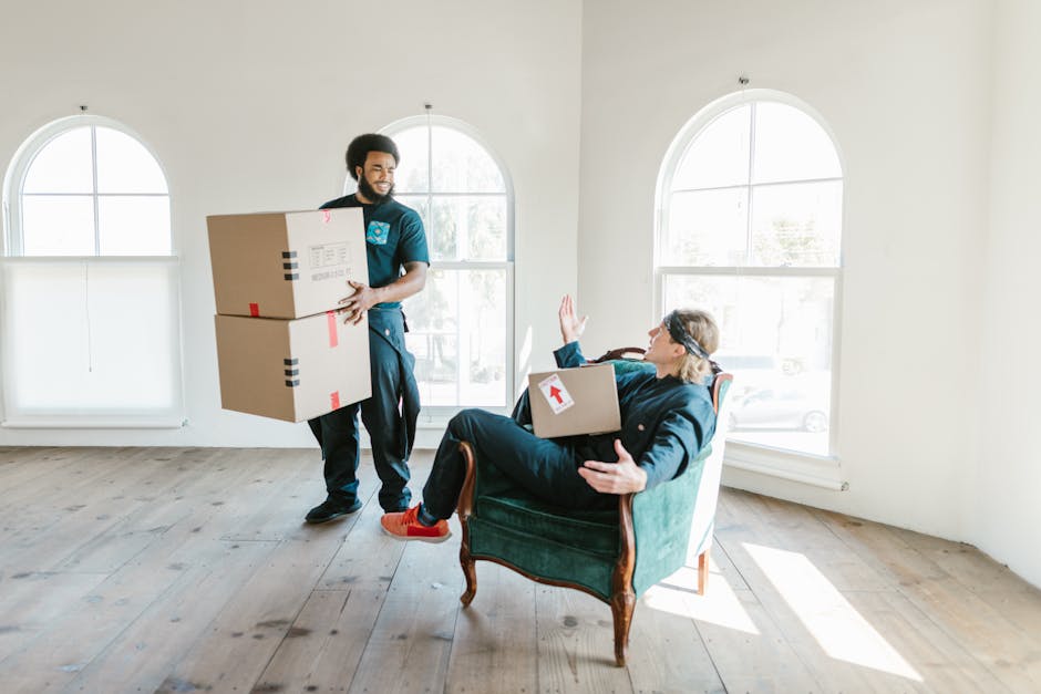A close-up view of a person wearing a dark blue shirt and matching blue work trousers, securing a large cardboard moving box that has a red and white caution label indicating 'This Side Up' with a dow