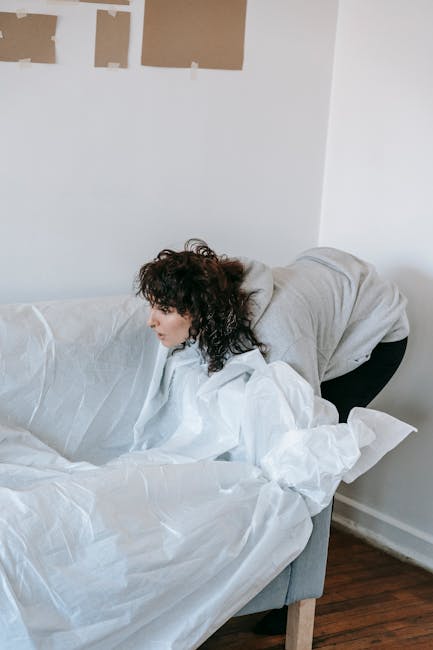 A professional removal team from Lewisham Removals is shown inside a well-lit, minimally decorated room with white walls and large, arched windows revealing a view outside. One mover, standing, is car