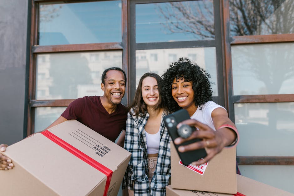 Three diverse individuals standing outdoors in front of a building with large glass windows, holding cardboard boxes sealed with red tape and smiling while taking a selfie with a mobile device. The person on the right, a woman with curly black hair and a white T-shirt, is extending her arm to hold the phone, capturing her friends next to her. The person on the left is a man with short black hair, wearing a maroon T-shirt, and the individual in the middle is a woman with straight brown hair dressed in a plaid shirt. The boxes appear to be packed with household items, suggesting they are in the process of a home relocation or moving activity. The scene is well-lit with natural daylight, indicating a positive moment during packing or moving logistics, relevant to the services of Lewisham Removals, a company specializing in removals and moving services, as supported by the context of the Lewisham station moving guide.